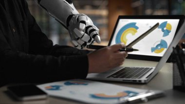 Pürüzsüz bir robot kol modern bir çalışma alanında bir insanla birlikte çalışır, bir dizüstü bilgisayarda dijital grafikler ve analizler gösterir, yenilik ve teknoloji entegrasyonunu sembolize eder.