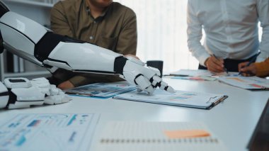 İnsan iş arkadaşlarıyla ortak bir toplantıya katılan bir robot kolu modern ofis masasındaki çizelge ve belgelere işaret ederek, takım çalışması sergiliyor.