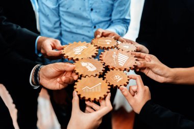 Çeşitli iş konseptlerini sembolize eden ahşap dişliler taşıyan, modern ofis ortamında takım çalışmalarını ve stratejik planlamayı yansıtan çeşitli profesyonellerden oluşan bir grup işbirliği yapıyor. Dostluk