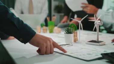 Sürdürülebilir enerji projelerine odaklanan çeşitli profesyonellerin yer aldığı işbirlikçi bir toplantı sahnesi. Eller modern ofis alanındaki tablolar ve verilerle etkileşir. SACTR