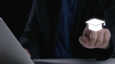 Mezuniyet şapkası ve çeşitli simgeler dijital çağda eğitim, bilgi ve profesyonel büyümeyi sembolize ederken bir kişi dizüstü bilgisayarla meşgul olur. Dörtgen