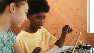 Afrika 'lı bir erkek ve beyaz bir kızın günlük giysiler içinde BTMM teknoloji sınıfında dizüstü bilgisayar ve elektronik ekipmanlarla kod yazma ya da programlama sisteminde birlikte çalıştıklarını gösteren site görüntüsü. Öğretim