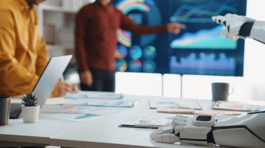 Modern bir ofiste yapılan bir iş toplantısında veri analizine yardımcı olan bir robot el sergileniyor. Ekip üyeleri yenilik ve işbirliğini yansıtan çizelge ve grafikleri araştırıyorlar. Noogenesis