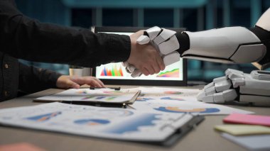 İnsan eli modern bir ofiste, teknoloji ve insan zekası arasındaki ortaklığı gösteren bir robot kolunu sıkıyor. Noogenesis