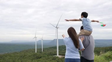 İlerici mutlu aile kavramı rüzgâr türbini çiftliğinde eğleniyor. Rüzgar türbini jeneratörüyle gelen elektrik jeneratörü kırsal bölgede tepe ve ufukta dağ var..