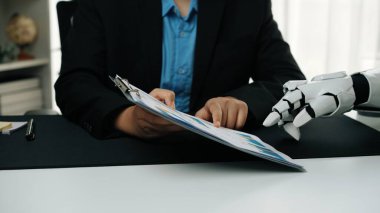 Bir insan eli, modern ofis ortamında robotik bir elin yardımıyla finansal belgeleri inceleyerek şirket dünyasındaki teknolojik gelişmeleri vurguluyor. Noogenesis