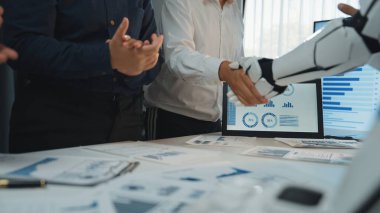 Modern bir ofiste, insan zekası ve teknoloji arasındaki sinerjiyi vurgulayan, insansı bir robotla el sıkışan iş dünyasından profesyonellerin dinamik bir sahnesi. Noogenesis 'in geleceği başarılı.