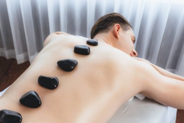 Lüks tatil köyündeki kaplıcada sıcak taş masajı. Gün ışığı dinginliği ortamı, mutlu erkek müşteri bazalt taşı masajının keyfini çıkarıyor. Rahatlatıcı bir sıcaklıkla vücudun üzerinde süzülüyor. Sessiz.