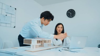 Profesyonel Asyalı erkek mimar ev modelinin uzunluğunu ölçmek için cetvel kullanırken, beyaz tenli genç meslektaşı ev modeliyle toplantı masasındaki verileri analiz etmek için dizüstü bilgisayar kullanıyor. Tertemiz..