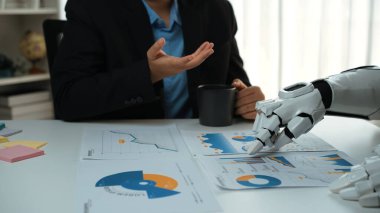 Bir insan ve robotik kol, çalışma masasındaki çizelge ve veri raporlarını analiz ederek, iş yerinde teknolojinin entegrasyonunu vurguluyor. Noogenesis