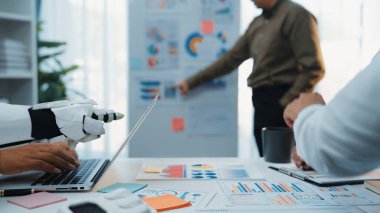 Robot kolun yanında dizüstü bilgisayar kullanan bir insan ofiste işbirliği gösterir. Veri tabloları modern bir iş yeri sunar. Teknoloji entegrasyonu ve takım çalışması. Noogenesis
