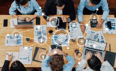 Çeşitli bir iş ekibi, veri bağlantıları ve içgörülerini gösteren dijital grafiklerle geliştirilmiş dizüstü bilgisayarlar ve belgeler kullanarak bir masa etrafında dinamik bir işbirliğine girişiyor. Dostluk