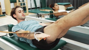 Bir grup müşteri, yenilikçi bir makinede bacak yükseltme işlemi yapıyor. Pilates partneri reformcu pozu veriyor ve egzersiz sırasında dengeyi geliştirmek ve koordinasyonu arttırmak için kayış takıyor. Alışkanlık.