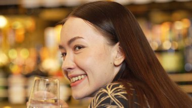 Yakın plan kadın içki tezgahı sahnesinde arkadaşlarıyla konuşurken elinde cin tonik bardağıyla içki içiyor. Bokeh ışıklandırma arka planındaki bulanık gece kulübünde güzel bir yüz. Vinosity.