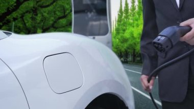 İlerici işadamı yeşil ormanın yakınındaki şarj noktasından elektrikli aracını şarj etmek için duruyor. Gelecekteki ortamı korumak için sürdürülebilir ve temiz enerji tarafından sürülen EV otomobil.