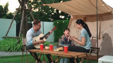 Baba gitar çalarken anne ve kız birlikte kampta oturuyorlar. Ebeveynler, genç nesil çapraz kuşak farklılıklarıyla zaman geçirmek için açık hava kamp aktivitelerini kullanıyorlar. Uyumsuzluk.