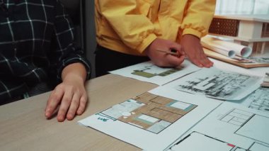 İki birey, şık bir çalışma alanındaki ahşap bir masanın ayrıntılı mimari planlarını ve çizimlerini inceleyerek ortak bir tasarım sürecine giriştiler. SACTR