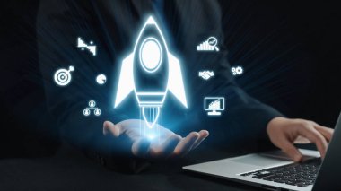 Bir iş adamı dijital bir dizüstü bilgisayarla etkileşime giriyor, elinde parlayan bir roket ikonu sergiliyor, yenilik, büyüme ve girişimci ruhu sembolize ediyor. Dörtgen