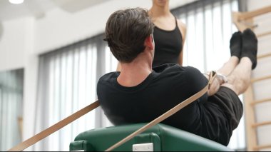 Müşteri, eğitmen desteğinin rehberliğinde kayışlarla Pilates reformatında egzersiz yapıyor. Sağlıklı bir adam pilates yapıyor ve üst vücut geliştirmek için Cadillac 'a asılıyor. Alışkanlık.