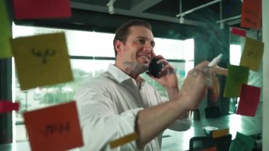 Profesyonel işadamı renkli yapışkan notlara yazılmış pazarlama stratejisi fikrine bakarken müşterilerine telefon ediyor. Proje yöneticisi duvardaki telefon analiz verileriyle konuşuyor. Yönlendirici.