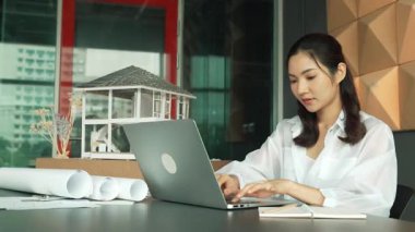 Genç ve güzel mimar mühendisin çatı malzemelerini kontrol etmesi ve ofisteki masaya yerleştirilmiş proje planı ve ev modeliyle dizüstü bilgisayarı kullanması. Yaratıcı iş tasarımı konsepti. Yönlendirici.