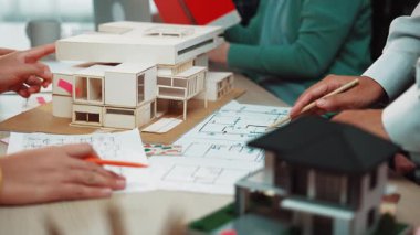 Modern bir tasarım stüdyosunda çalışan profesyonellerden oluşan bir ekip, mimari bir model ve planlara odaklanıyor. Nişanlı eller yaratıcı planlama sürecini gösteriyor. SACTR