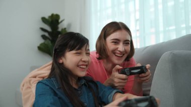 Annem dizüstü bilgisayar kullanarak kanepeye uzanırken, mutlu kız oyun oynuyor ve oturuyor. Beyaz bir ebeveyn ve çekici bir kız, elinde joystick ile birlikte vakit geçirir ve maç kazanmaya odaklanırlar. Pedagoji.