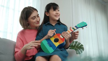 Kafkasyalı bir anne evde akustik müzik öğretirken ukulele çalan mutlu kız. Sevimli çocuk enstrüman öğreniyor. Kafkasyalı mutlu anne ve kız birlikte vakit geçiriyorlar. Pedagoji.