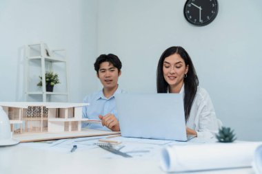 Profesyonel Asyalı erkek mimar ev modelinin uzunluğunu ölçmek için cetvel kullanırken, beyaz tenli genç meslektaşı ev modeliyle toplantı masasındaki verileri analiz etmek için dizüstü bilgisayar kullanıyor. Tertemiz..