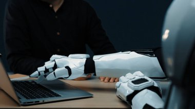 İnsan-robot etkileşiminin yakın plan görüntüsü, masadaki dizüstü bilgisayara uzanan bir robot kolu gösteriyor, teknolojinin ve işbirliğinin geleceğini yansıtıyor. Noogenesis