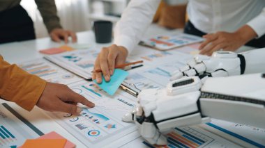 Bir insan ve bir robot, renkli yapışkan notlar ve detaylı belgelerle çevrili, modern ofis ortamında takım çalışması ve yenilik içeren bir proje üzerinde işbirliği yapıyorlar.