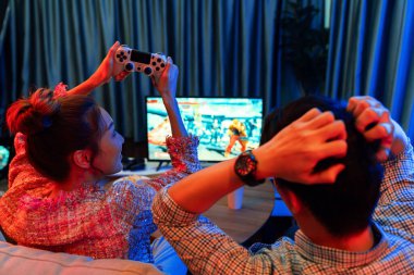 Joystickli birkaç oyuncu dövüş oyunu oynuyor, kadın oyuncu yarışması derecesi arka tarafta TV ekranında modern rahat evinde neon ışıklı oturma odasındaki koltukta oturuyor. İnfobahn.