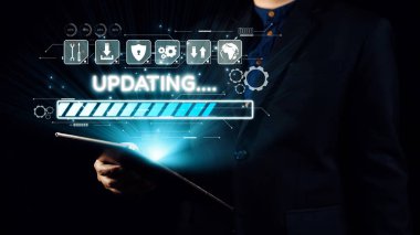 Koyu renk takım elbiseli bir iş adamı, bir şirket ortamında teknoloji ve yenilik temalarını gösteren, güncellemeli dijital arayüzü gösteren bir tablet tutuyor. Dörtgen