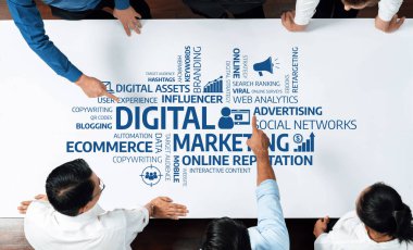 Bir grup iş adamı, anahtar terimleri ve kavramları etkin beyin fırtınası için bir masada görsel olarak sergilenen dijital pazarlama stratejilerini işbirliği içinde tartışıyor. Dostluk