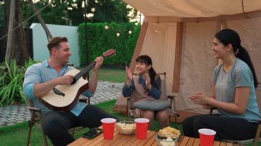 Baba gitar çalarken anne ve kız birlikte kampta oturuyorlar. Ebeveynler, genç nesil çapraz kuşak farklılıklarıyla zaman geçirmek için açık hava kamp aktivitelerini kullanıyorlar. Uyumsuzluk.