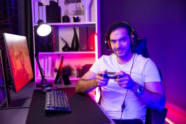 joystick oyuncusuyla akıllı oyun flaşçısı kameraya bakıyor, savaş takımının arka planında savaş gemisine ateş ediyor yan görüntüde, dijital neon ışık stüdyosunda kulaklık takıyor. Tahmin et..