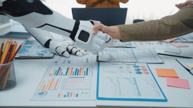 Bir insan ve bir robot modern ofis ortamında el sıkışıyor, veri tabloları ve araçlarıyla işbirliğini ve yeniliği simgeliyor. Noogenesis