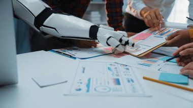 Bir robot kolu, ortak bir toplantı sırasında bir insan ekibine yardım eder, ofis masasındaki veri raporlarını ve çizelgeleri analiz eder, iş dünyasındaki takım çalışmalarının geleceğini vurgular. Noogenesis