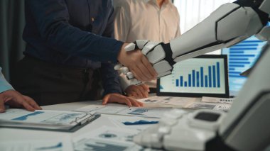 Bir insan ve bir robot modern ofis ortamında el sıkışıyor, iş dünyasındaki takım çalışmasını ve yeniliği simgeliyor, ekranlarda finansal analizler gösteriliyor. Noogenesis