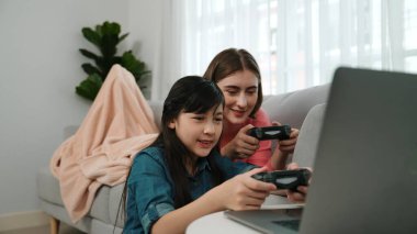 Annem dizüstü bilgisayar kullanarak kanepeye uzanırken, mutlu kız oyun oynuyor ve oturuyor. Beyaz bir ebeveyn ve çekici bir kız, elinde joystick ile birlikte vakit geçirir ve maç kazanmaya odaklanırlar. Pedagoji.