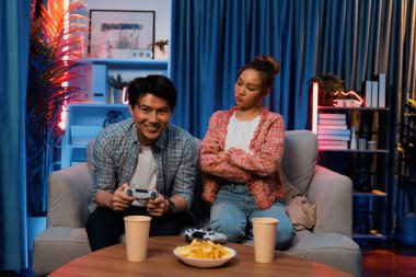 Mutlu çiftin oturduğu kanepe joysticklerle video oyunu oynuyor, meydan okuyan gülümseyen adam, evdeki modern neon ışıklı oturma odasında kazanmak için yumruğunu kaldırıyor. İnfobahn.