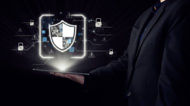 Koyu renk elbiseli bir profesyonel, bugün dijital ortamda siber güvenlik ve veri korumanın kritik önemini temsil eden parlayan bir kalkanın bulunduğu bir tablet sergiliyor. Dörtgen