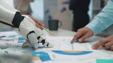 Robot bir kol, modern çalışma alanlarındaki teknoloji ve takım çalışmalarının karışımını gösteren bir iş toplantısı sırasında çizelgeleri analiz etmede profesyonellere yardımcı olur. Noogenesis