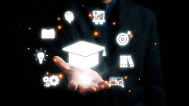 Çeşitli eğitim simgeleriyle çevrili bir mezuniyet şapkası sembolüne uzanan bir el, karanlık bir arkaplanda bilgi, gelişme ve akademik başarıyı sembolize ediyor. Dörtgen