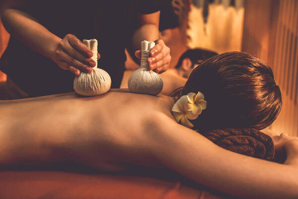 Hot herbal ball spa massage body treatment, masseur gently compresses herb bag on couple customer body. Serenity of aromatherapy recreation in warm lighting of candles at spa salon. Quiescent