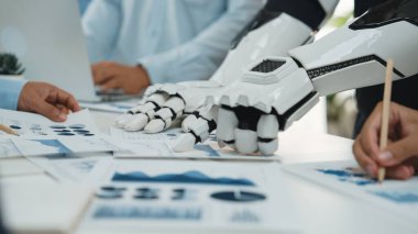 Robot bir kol ve insan elini, şık bir ofis ortamında veri analizi üzerinde çalışırken gösteren çarpıcı bir görüntü, teknolojinin iş birliğindeki rolünü vurguluyor. Noogenesis