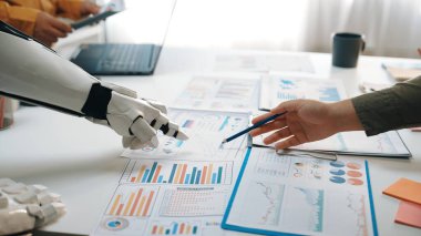 Bir insan ve robot, modern bir ofis ortamında finansal tablolar ve raporlarla veri analizi yaparak iş birliği yapıyor ve yenilikler sergiliyor.