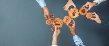 Çeşitli iş simgeleriyle dişlileri bir arada tutmak için birleşen çoklu ellerin geniş açılı bir görünümü, iş yerindeki işbirliğini, takım çalışmasını ve yenilikçi gelişimi sembolize ediyor. Dostluk