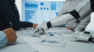 Dinamik bir iş toplantısı sahnesi. Bir insan ve bir robot kol, modern çalışma alanlarında teknolojinin entegrasyonunu gösteren veri raporlarını işbirliği içinde analiz ediyor. Noogenesis