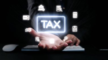 Mali sembollerle çevrili, karanlık bir ortamda parlayan bir vergi simgesini gösteren bir el uzanıyor. Finansal hizmetleri, danışmanlık ve teknolojiyi temsil ediyor. Dörtgen